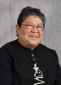 A person wearing glasses and a black shirt with a white cross pendant, smiling against a gray textured background.