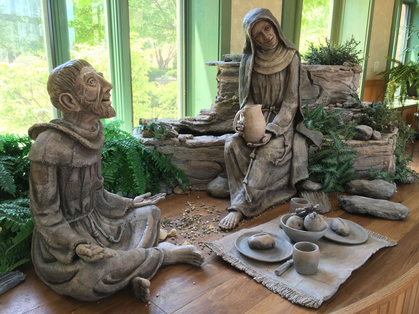Two stone-like figures, one seated and one kneeling, beside a small indoor fountain with plates and a cup on a cloth.