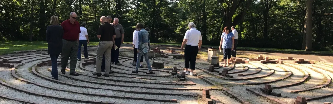 A group of people walk through a circular stone labyrinth outdoors on a sunny, wooded path.