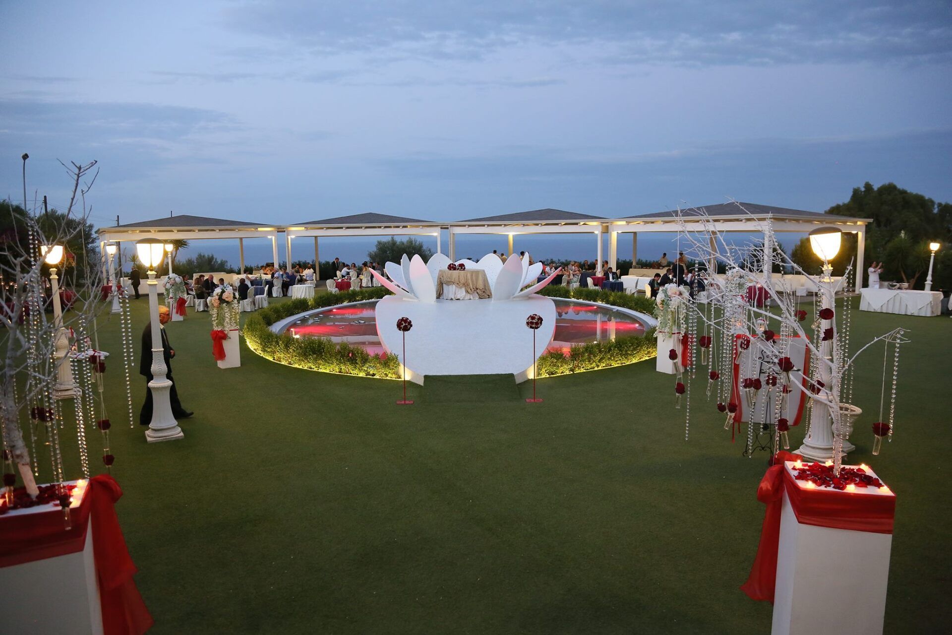 Location per matrimoni all'aperto decorata al crepuscolo con archi floreali, sedie bianche e un palco centrale su un prato verde.