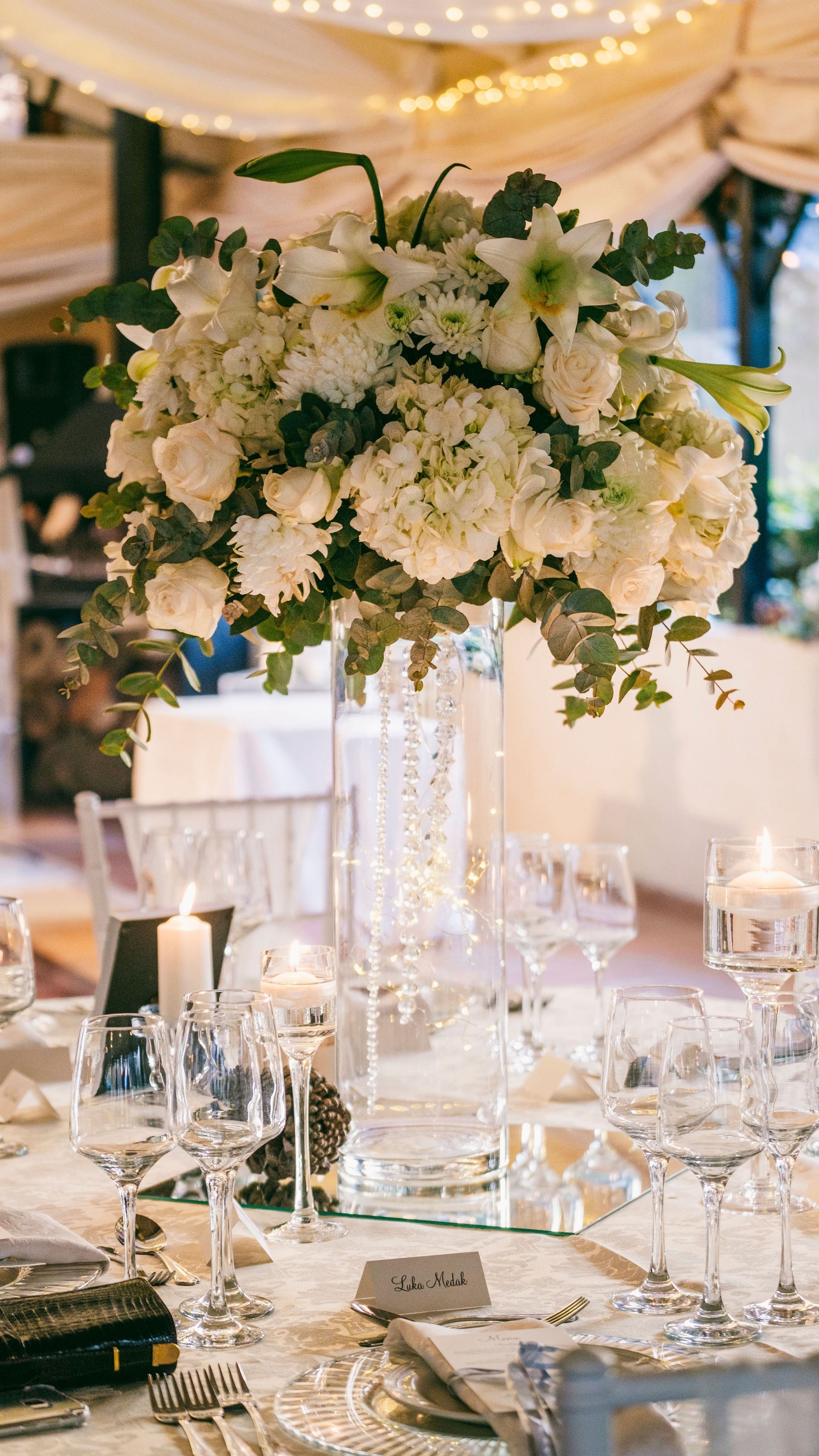 Elegante tavolo per il ricevimento di nozze con centrotavola floreale bianco, candele, bicchieri e dettagli dorati.