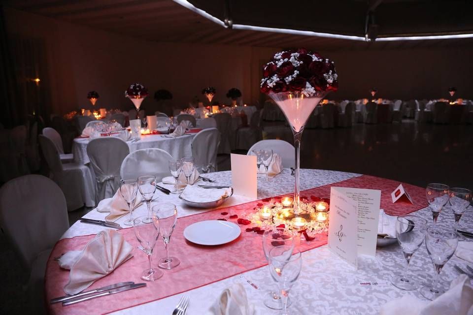 Elegante tavolo da banchetto con tovaglie bianche, tovaglioli piegati, candele e centrotavola floreale in una sala eventi illuminata da luci soffuse.