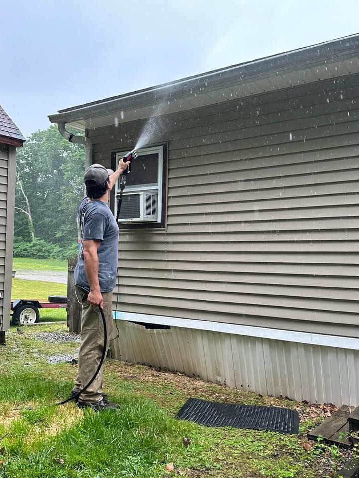 Man washing house siding with a hose outdoors. Beige siding, green grass, overcast sky.