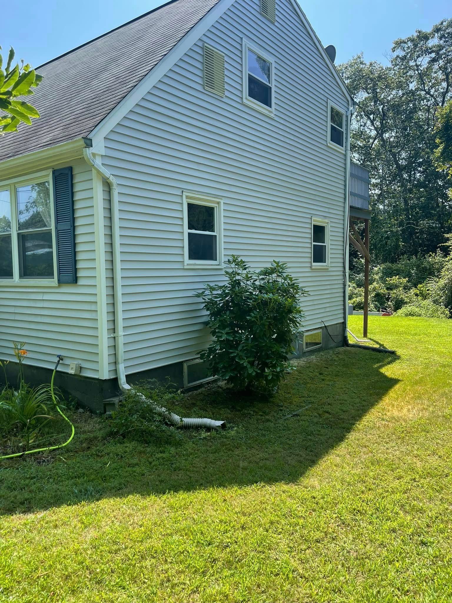Side view of a light blue house with a small bush in front. Green grass and trees surround it.