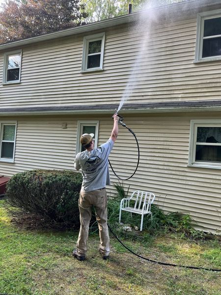Man power washing a light-colored house. Green grass, white chair, and windows visible.