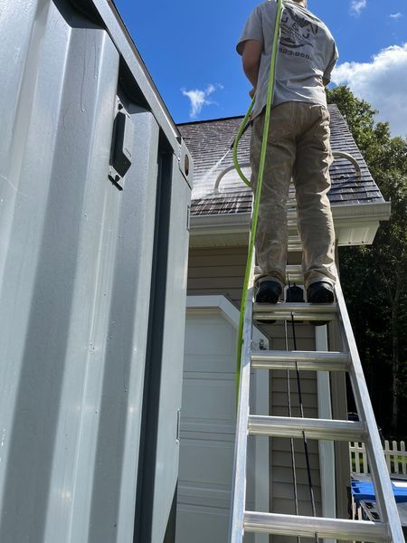 Person on a ladder, cleaning a house's roof. Green hose in hand, against a sunny sky.