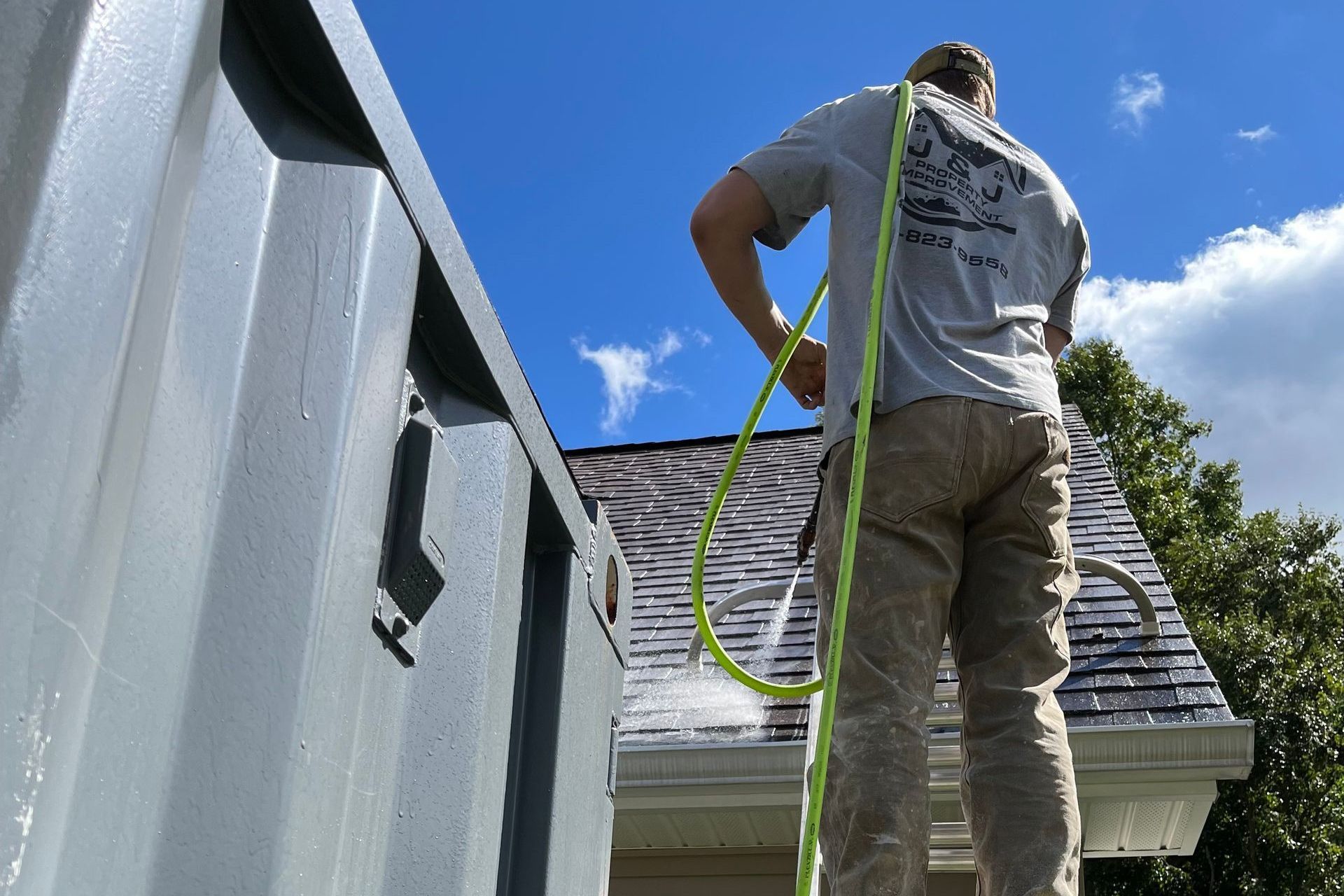 Person on a ladder spraying water on a house's roof and gutter on a sunny day.
