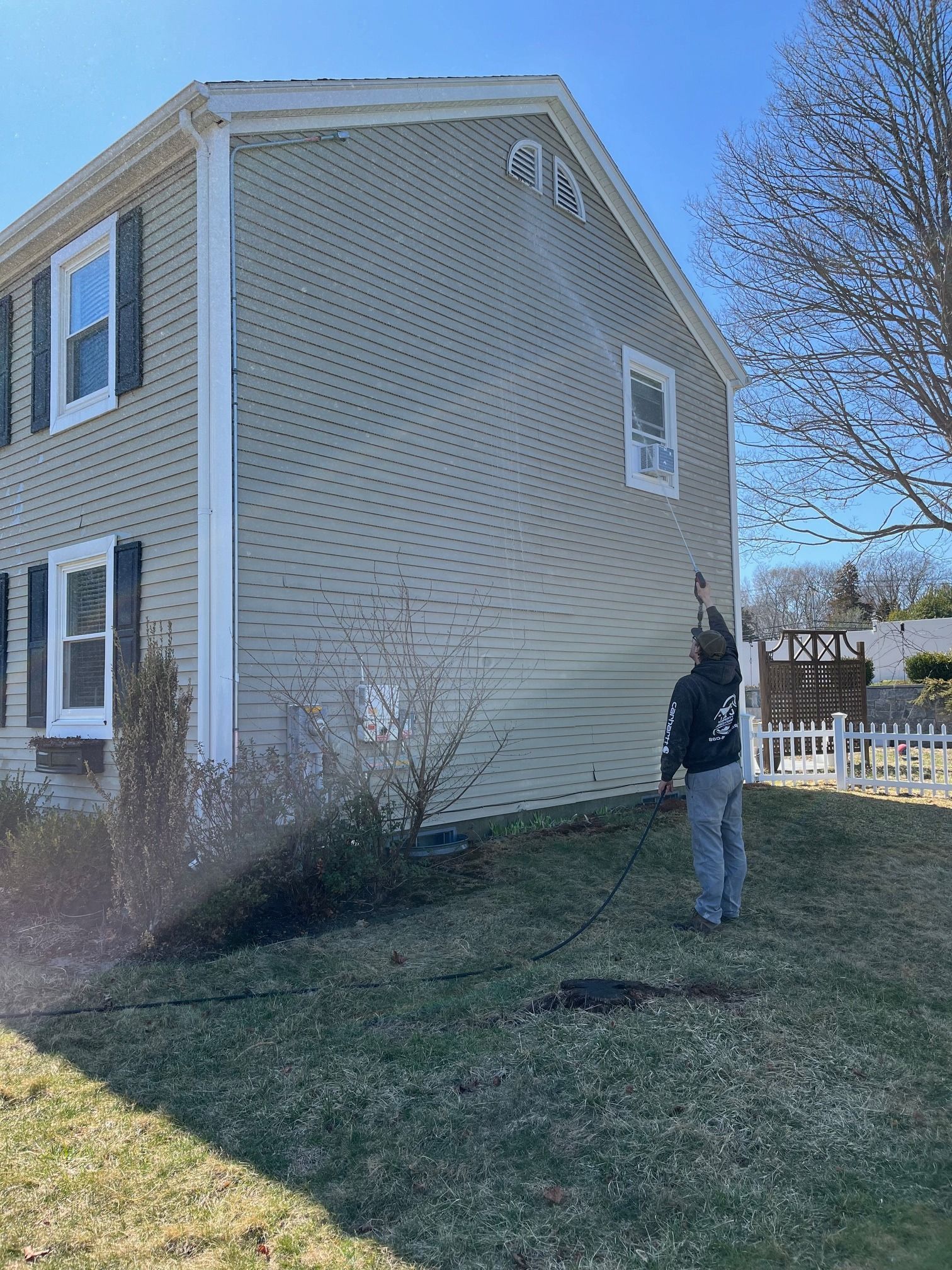 Person power washing the side of a two-story beige house with green lawn on a sunny day.
