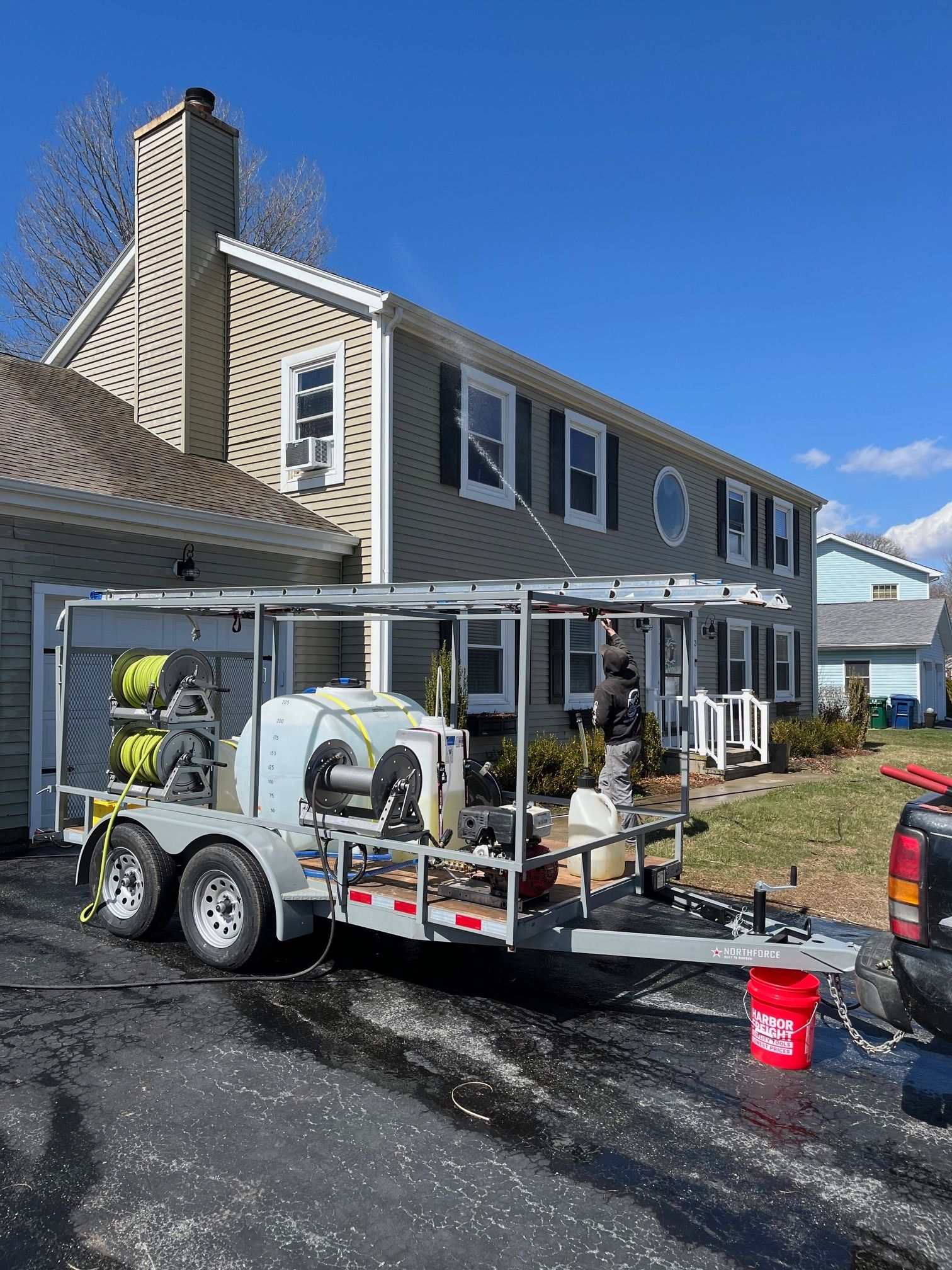 Pressure washing trailer in front of a house. A worker sprays the siding on a sunny day.