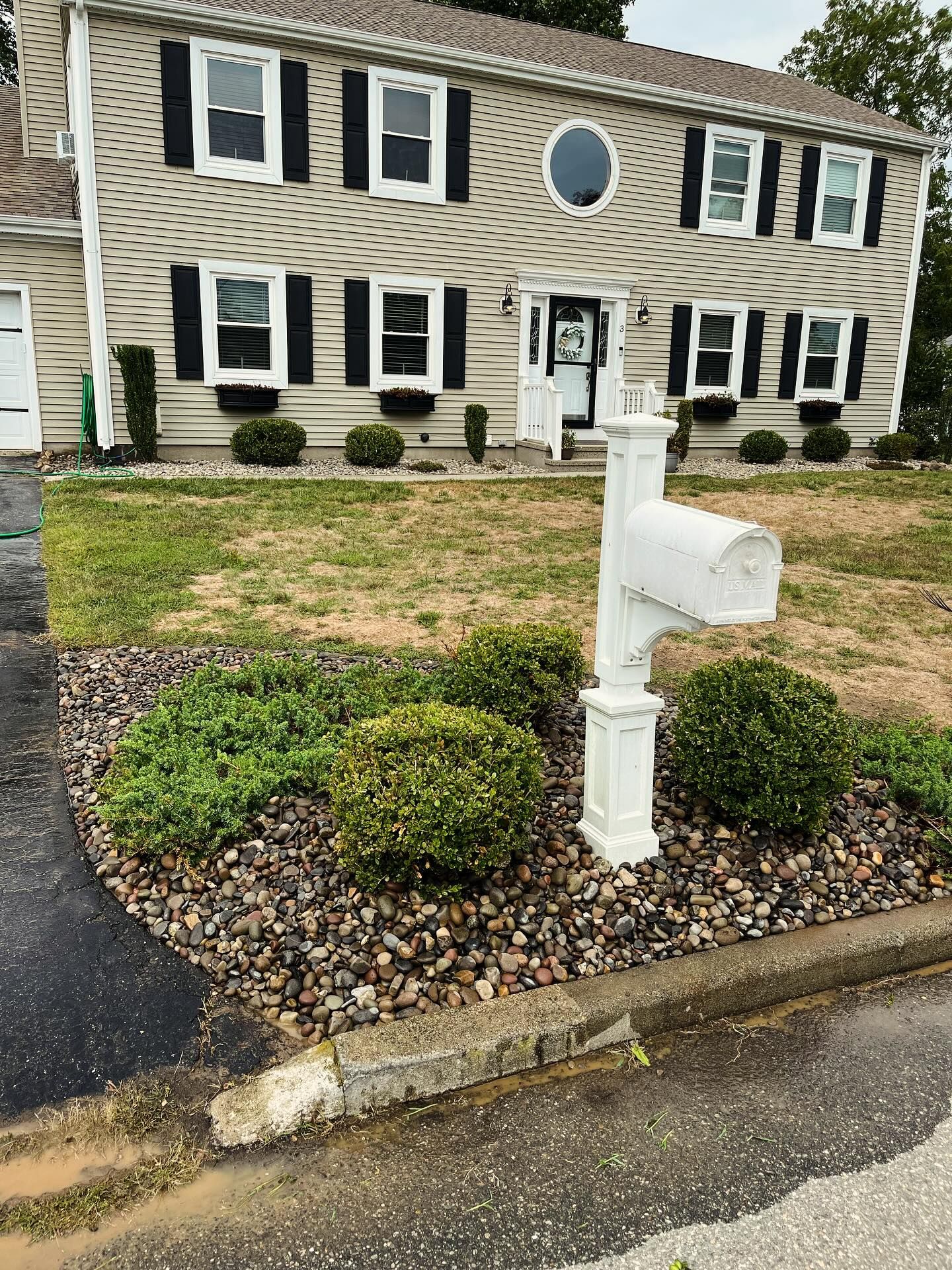 A two-story beige house with black shutters, a white mailbox, and a rock garden.