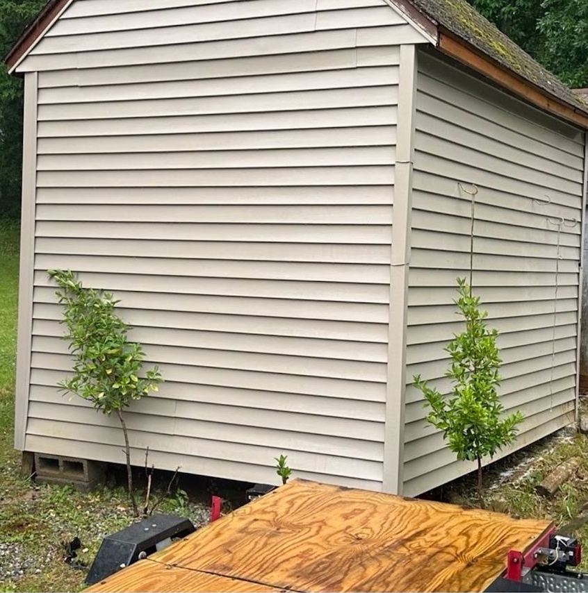 Tan shed with horizontal siding and two small trees in front, next to a wood-covered trailer.