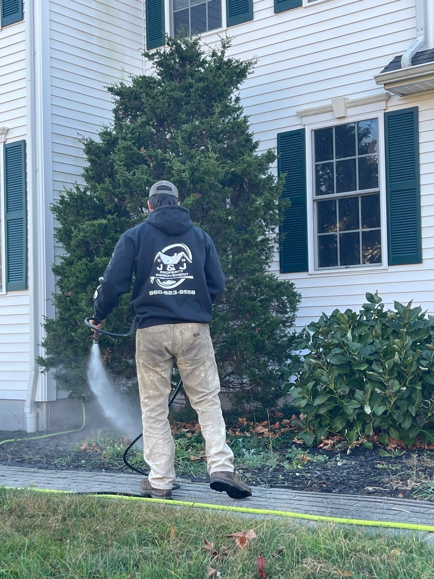 Person in gray hoodie sprays a hose near a white house with green shutters and a bush.