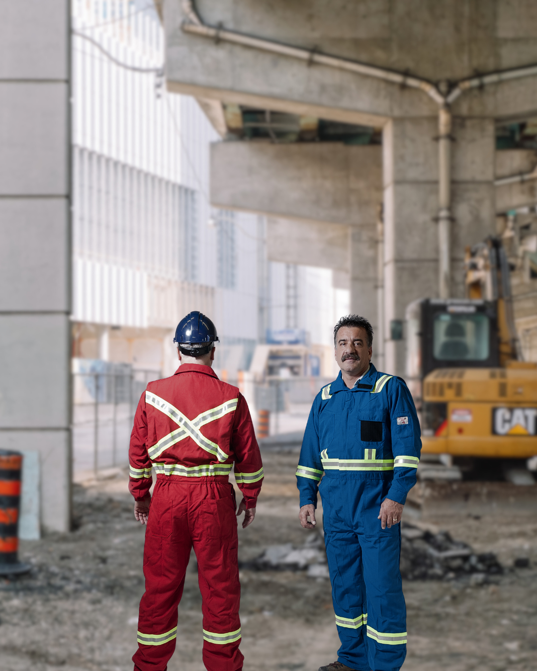 Two construction workers in coveralls stand under a bridge. One faces away, the other toward the camera.