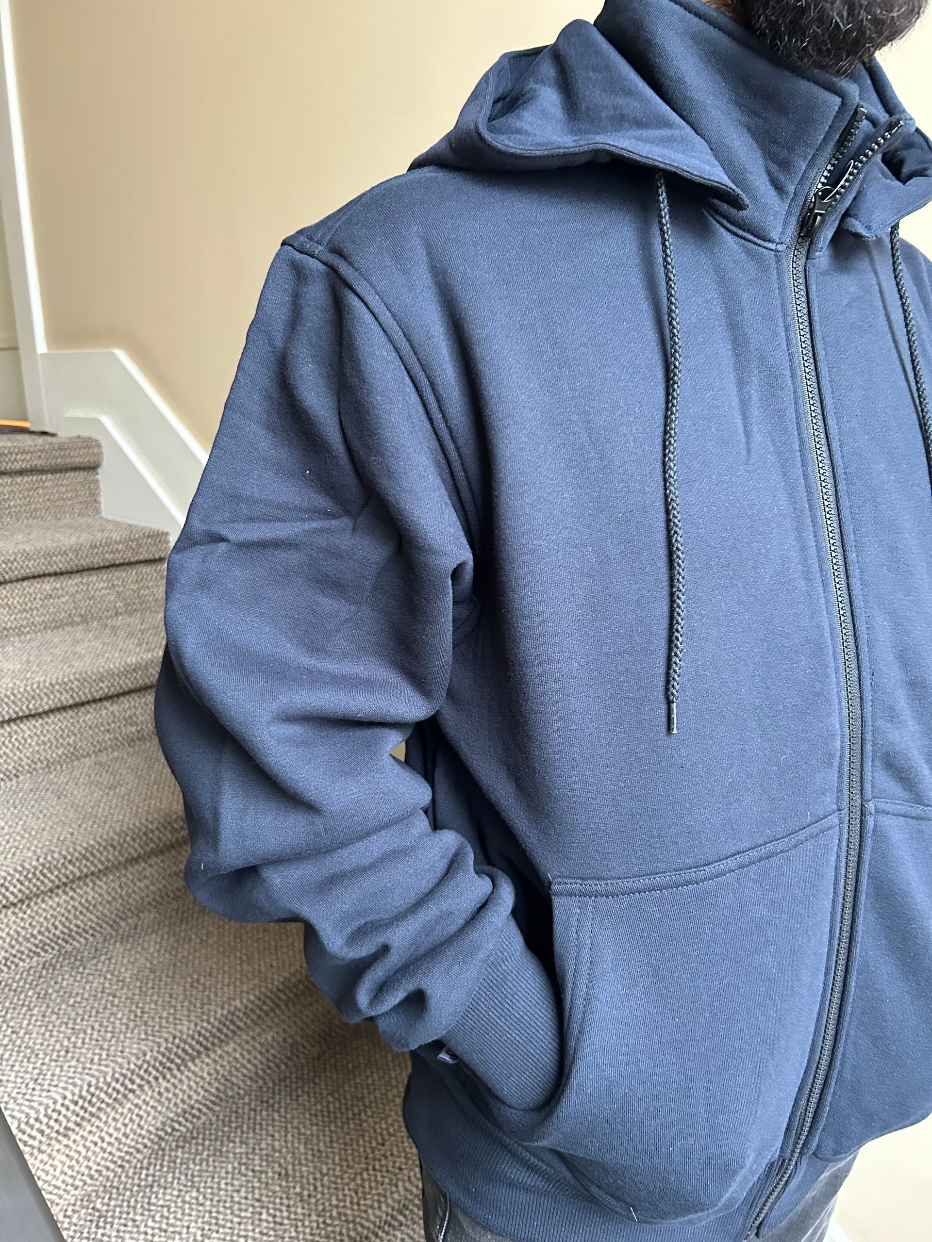 Person wearing a navy blue hooded zip-up sweatshirt with their hand in the pocket, standing near stairs.