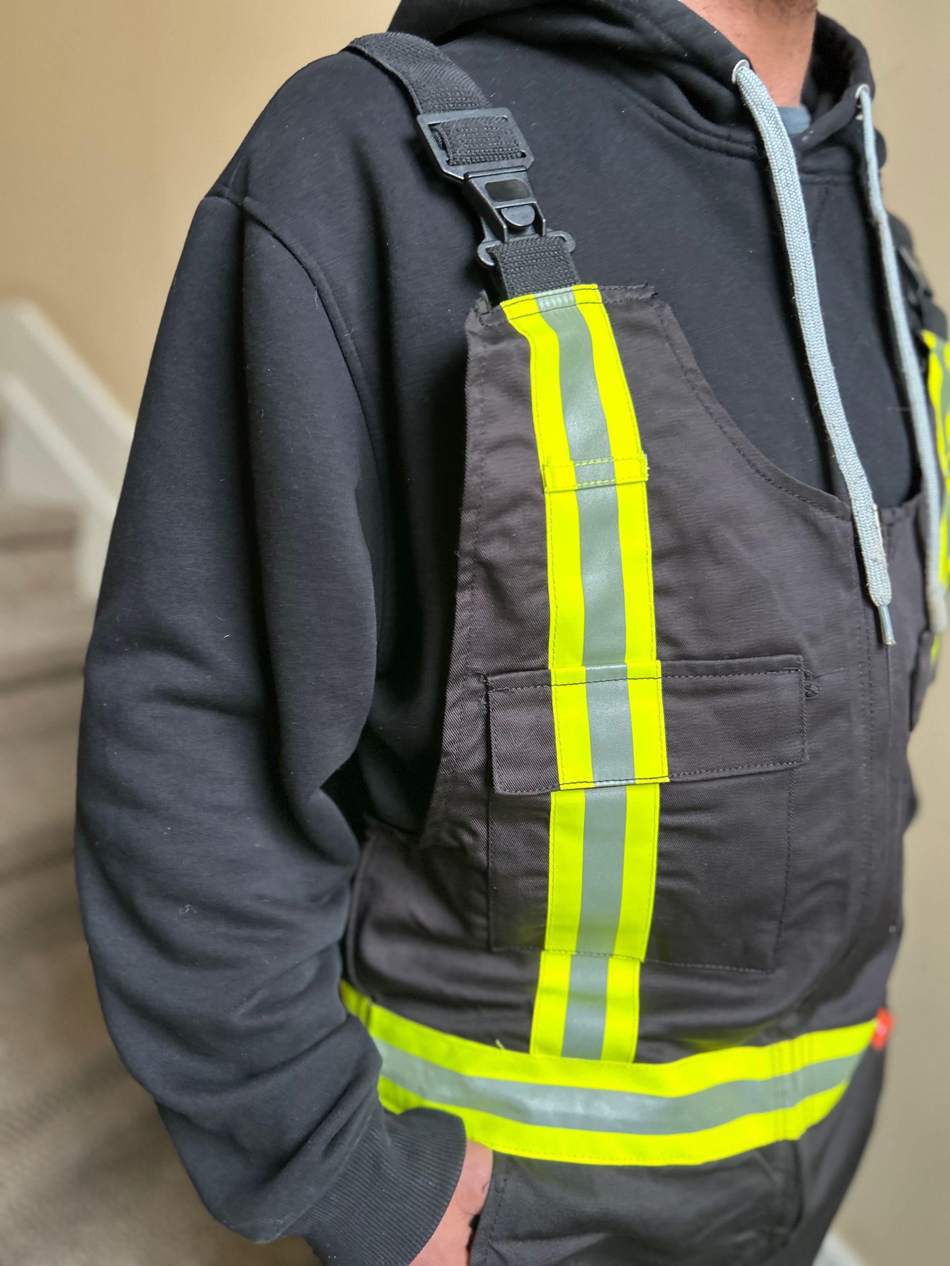 Person wearing black bib overalls with reflective yellow stripes over a black hoodie.
