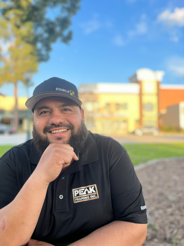 Man in black cap and polo shirt smiles, hand to chin, outdoors. Background: blurred trees and building.