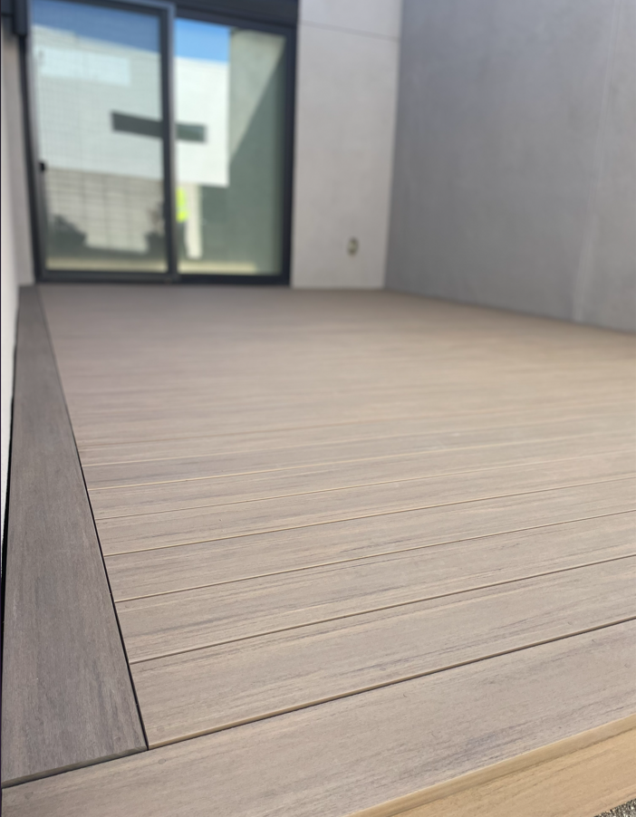 Wooden deck with a dark border, next to a sliding glass door and concrete wall.
