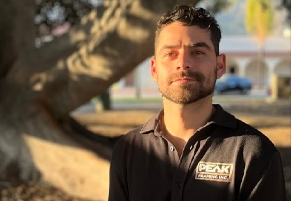 Man in black shirt with logo, stands outside under a tree.