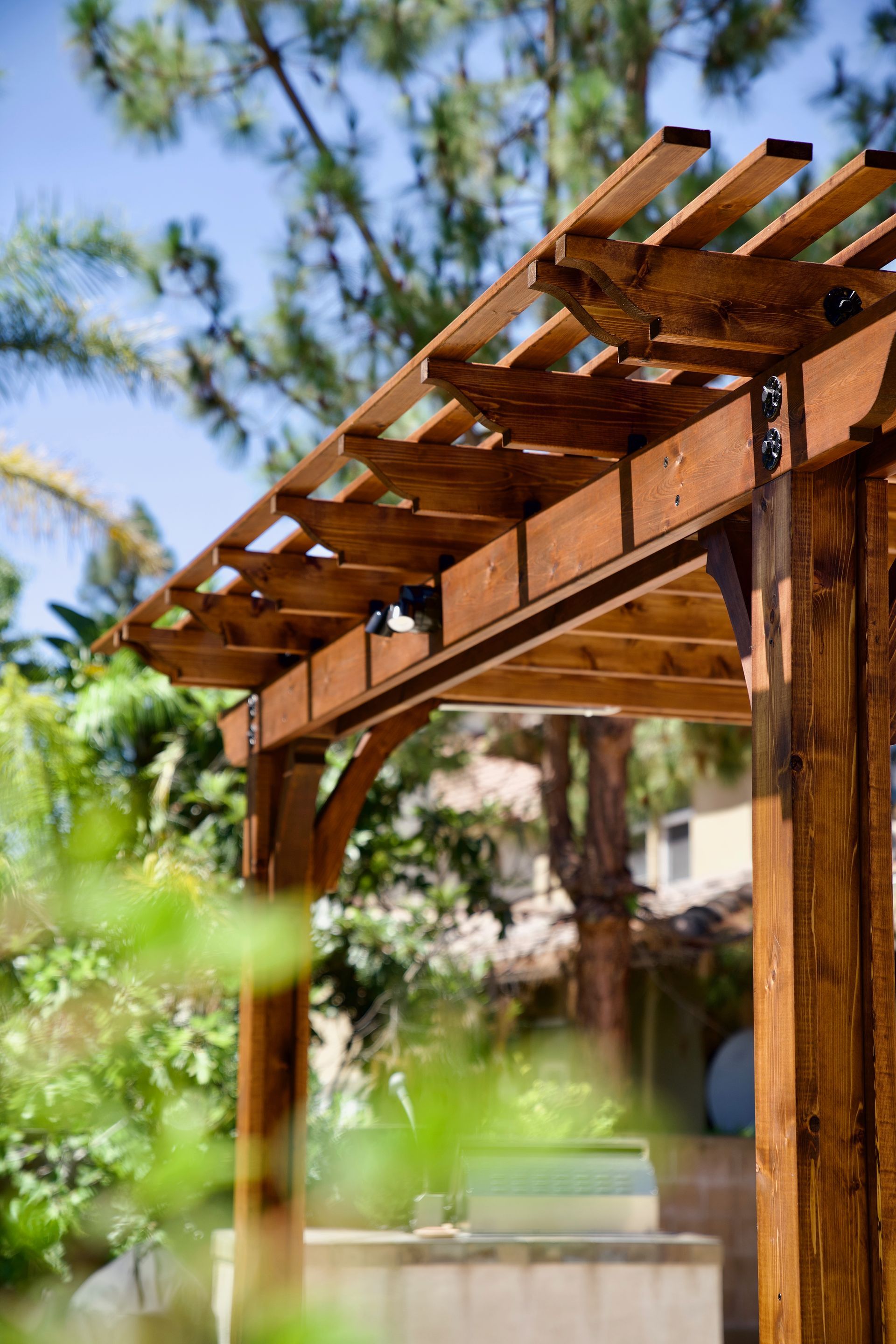 Wooden pergola with beams and slats, against a backdrop of trees and blue sky.