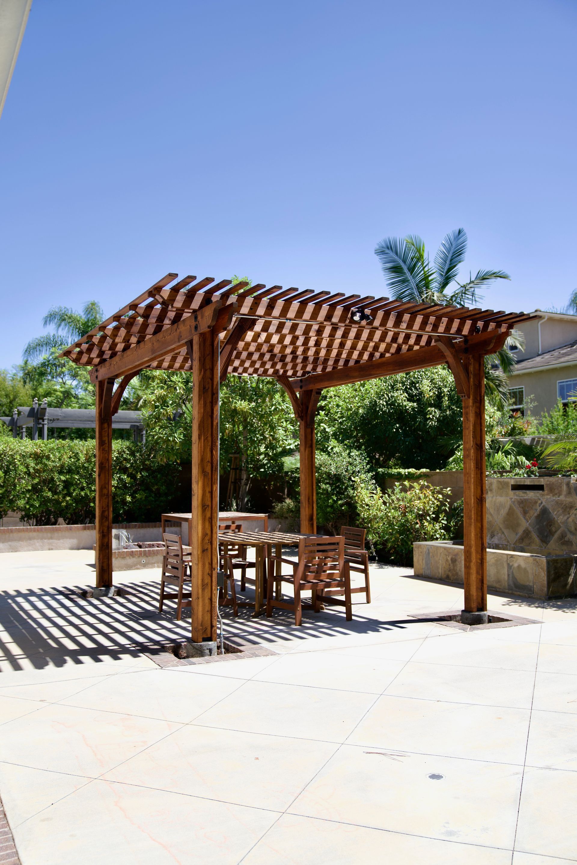 Wooden pergola over outdoor dining table, casting shadows on the patio, sunny day.