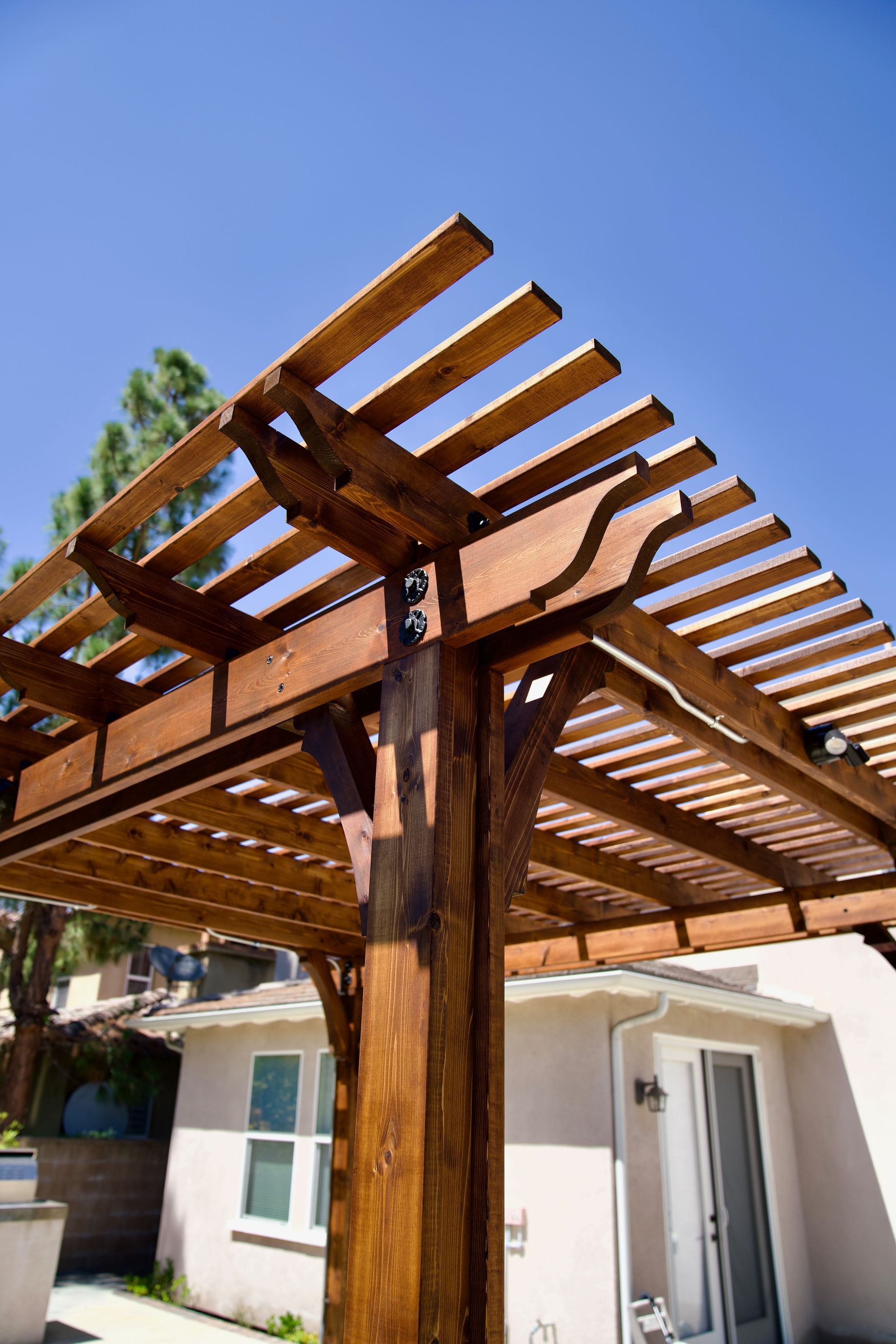 Wooden pergola in a sunny backyard, brown beams and rafters, blue sky.