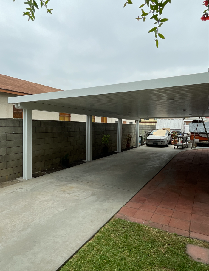 A covered carport with a concrete driveway. A white car and a trailer are parked inside. Overcast sky.