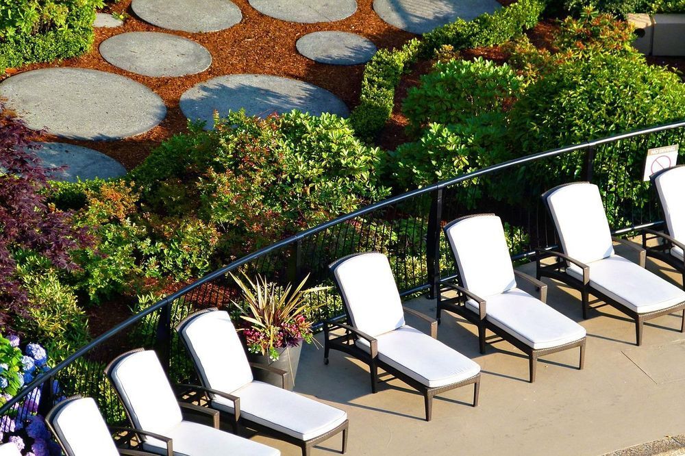 Lounge chairs on a patio with a garden in the background. Stone pathway, green shrubs, and a black metal railing.