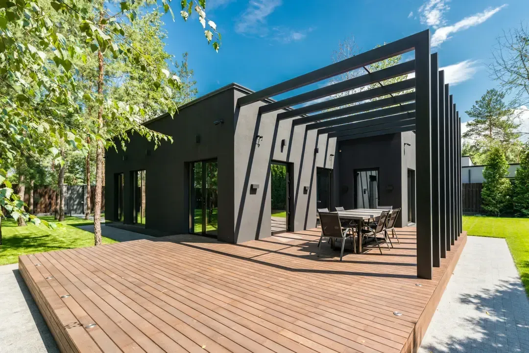 Modern black house with wooden deck and pergola, set on a green lawn with trees.
