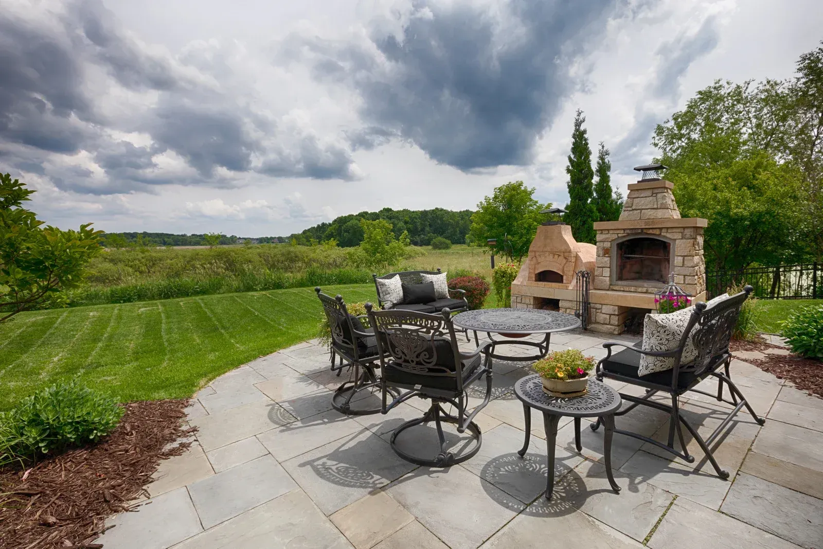 Outdoor patio with stone pizza oven, table, chairs, and field under cloudy sky.
