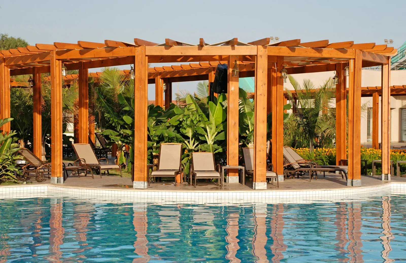 Poolside pergola with lounge chairs. Water reflects the wooden structure.