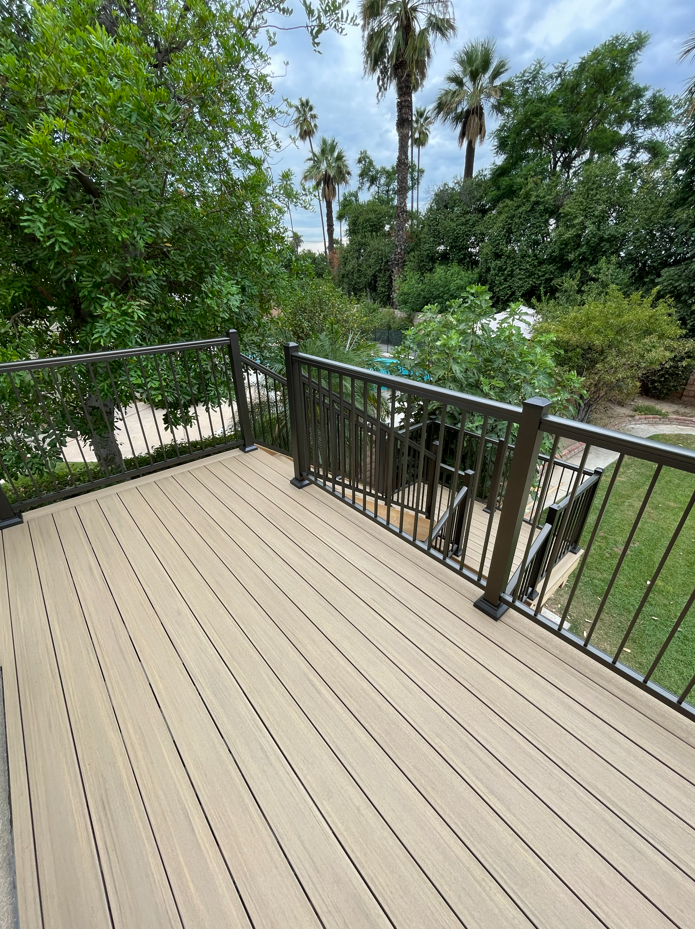 Deck with brown railing overlooks a green, leafy landscape with palm trees under a cloudy sky.