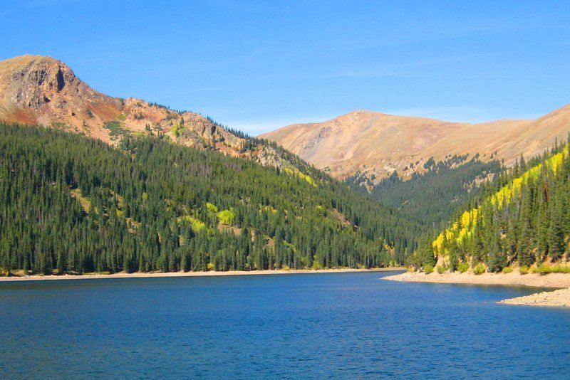 Blue lake nestled between green forested slopes and tan mountains under a clear blue sky.