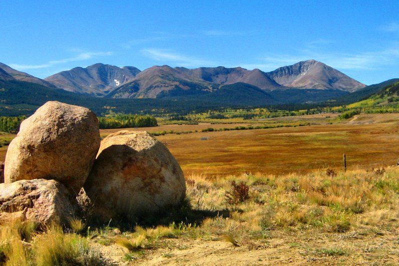 Large boulders in foreground, field of brown grass, mountains in background, clear blue sky.