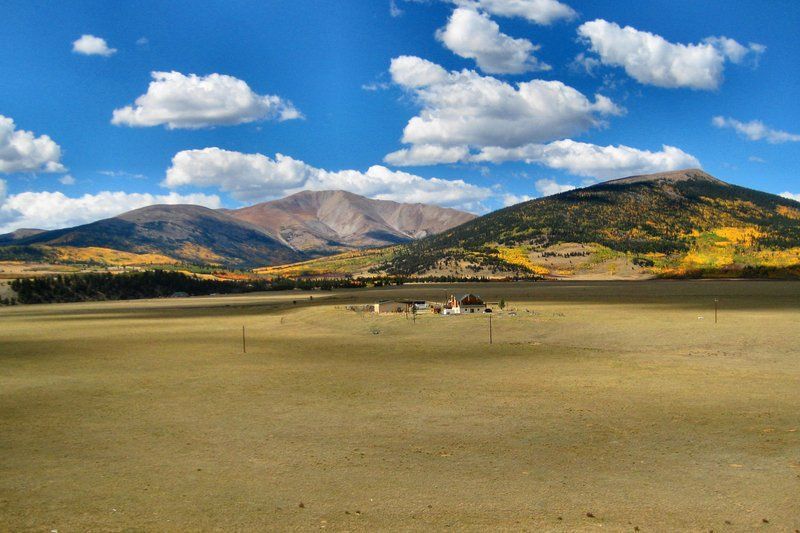 Mountains and field under a blue sky with fluffy clouds. Some fall colors on the mountain slopes.