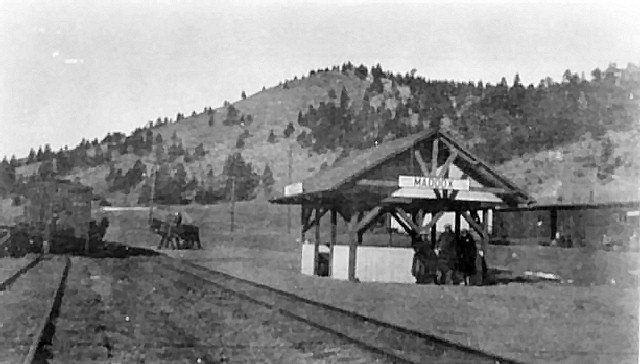 Vintage photo of a train depot. Tracks in foreground, riders under shelter. Hill in background.