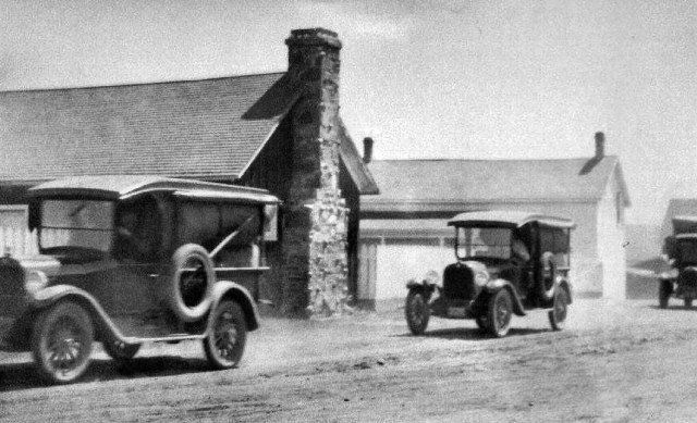 Vintage cars driving on a dirt road, passing a building with a stone chimney.