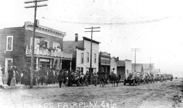 Parade in Fairplay, Colorado, with people on a street lined with buildings and a line of horse-drawn carriages.
