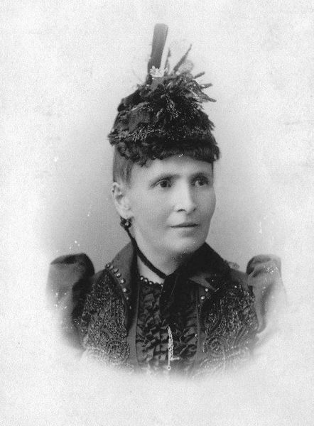 Woman wearing a decorated hat and dark coat, photographed from the chest up.