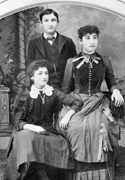 Three people posed for a portrait: a person standing in a suit, and two women in dresses, one seated.