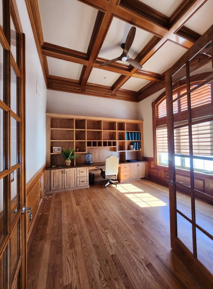 Wooden office with built-in shelves, desk, and ceiling beams. Sunlight streams in from windows.