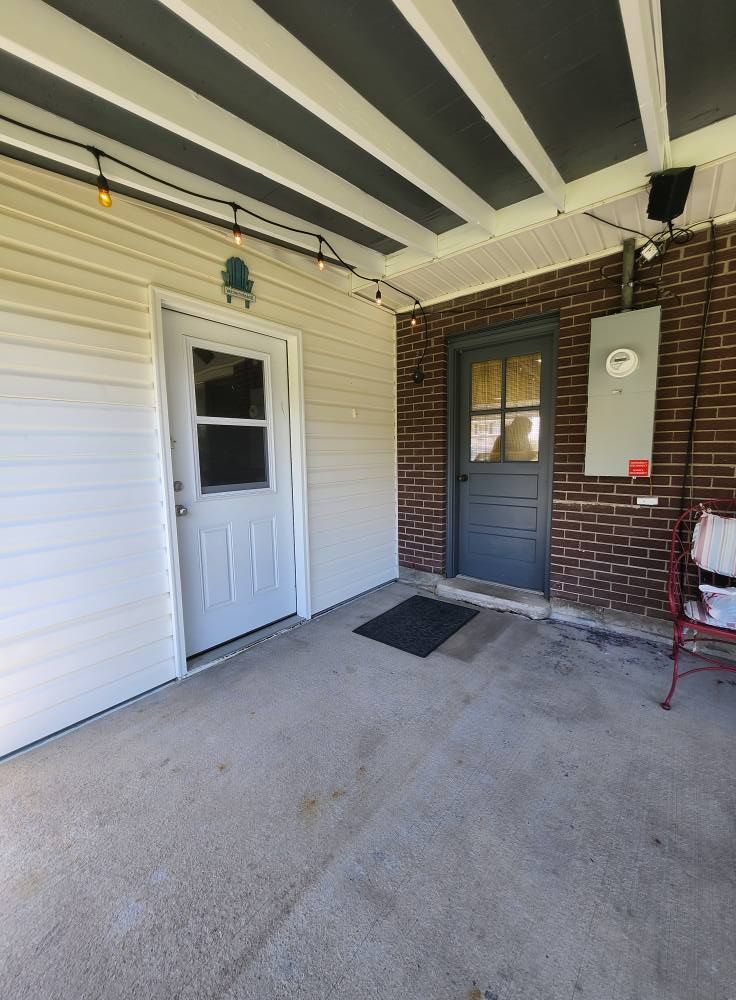 Covered patio with two doors, brick wall, and beige siding. A string of lights hangs overhead.