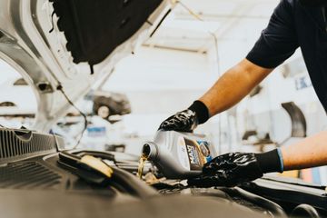 A person in work gloves pours motor oil from a bottle into a car engine at an auto repair shop.