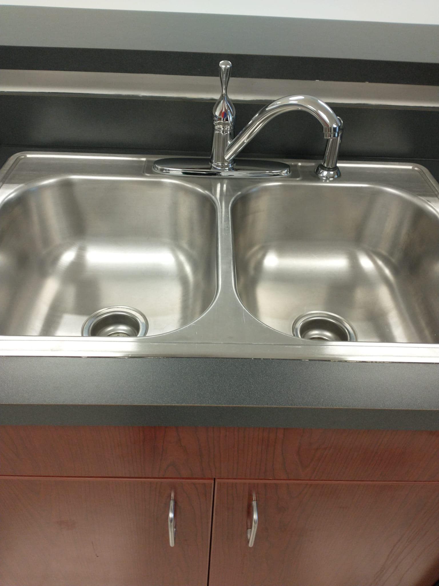Stainless steel double sink with chrome faucet, mounted in dark countertop above brown cabinets.
