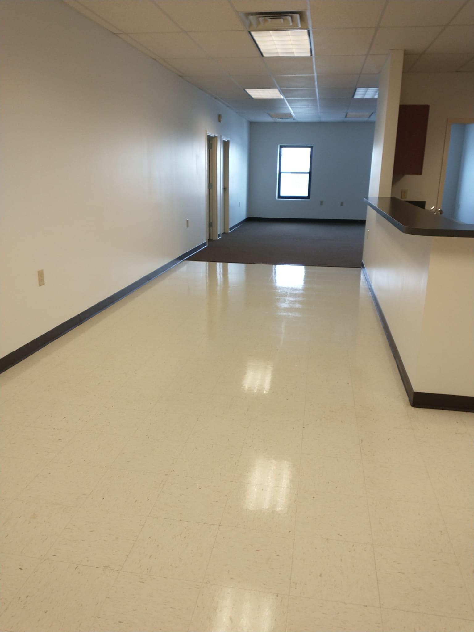 Empty office hallway with white walls and floor, leading to a dark brown carpeted area with a window.