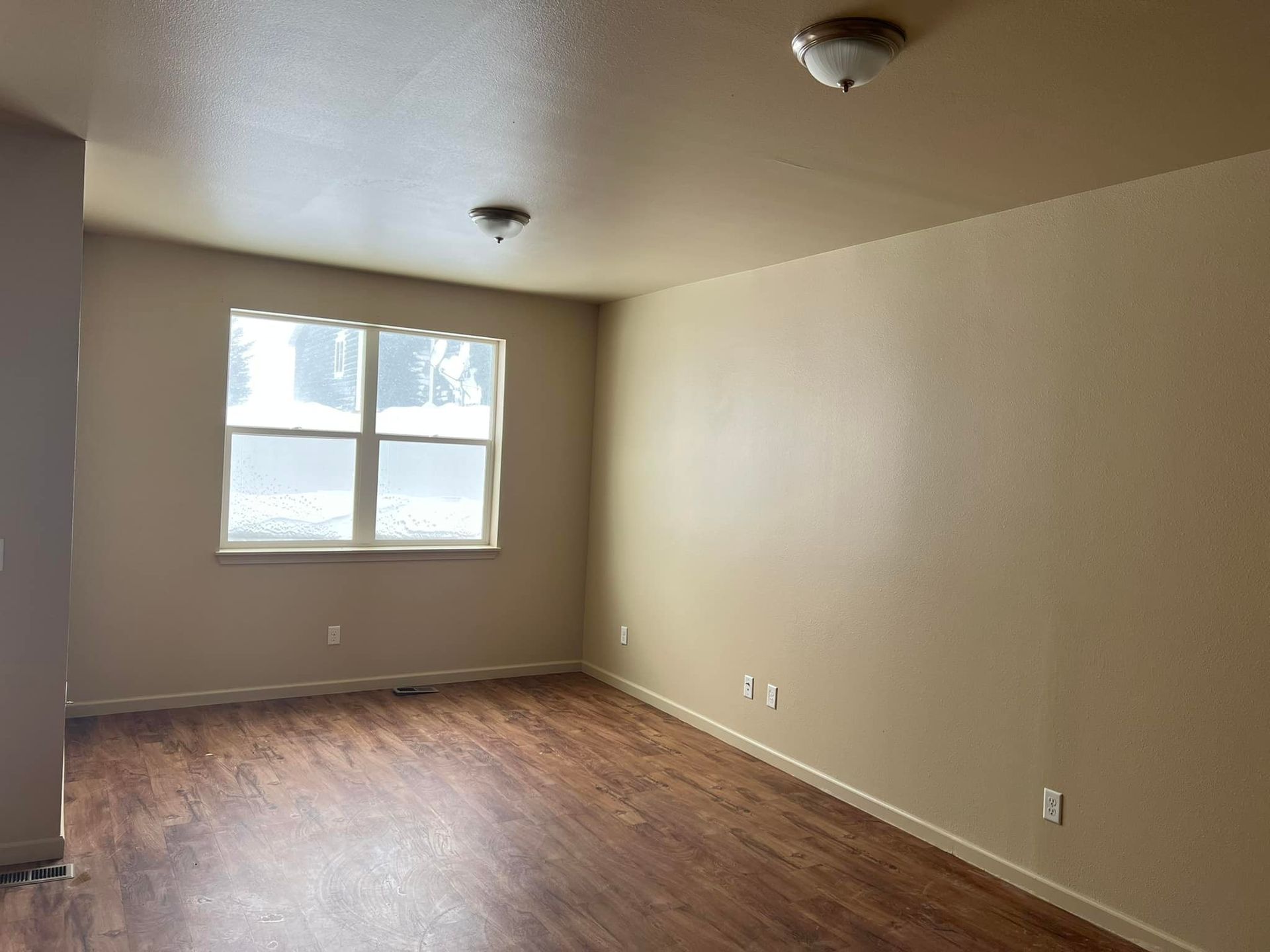 Empty room with tan walls, a window, and wood-look flooring. Two lights on the ceiling.