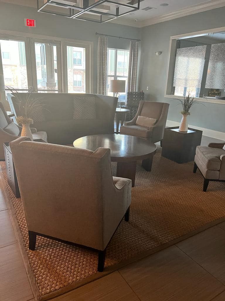 Living room with beige furniture, a rug, and a round table. Windows and a door are visible.