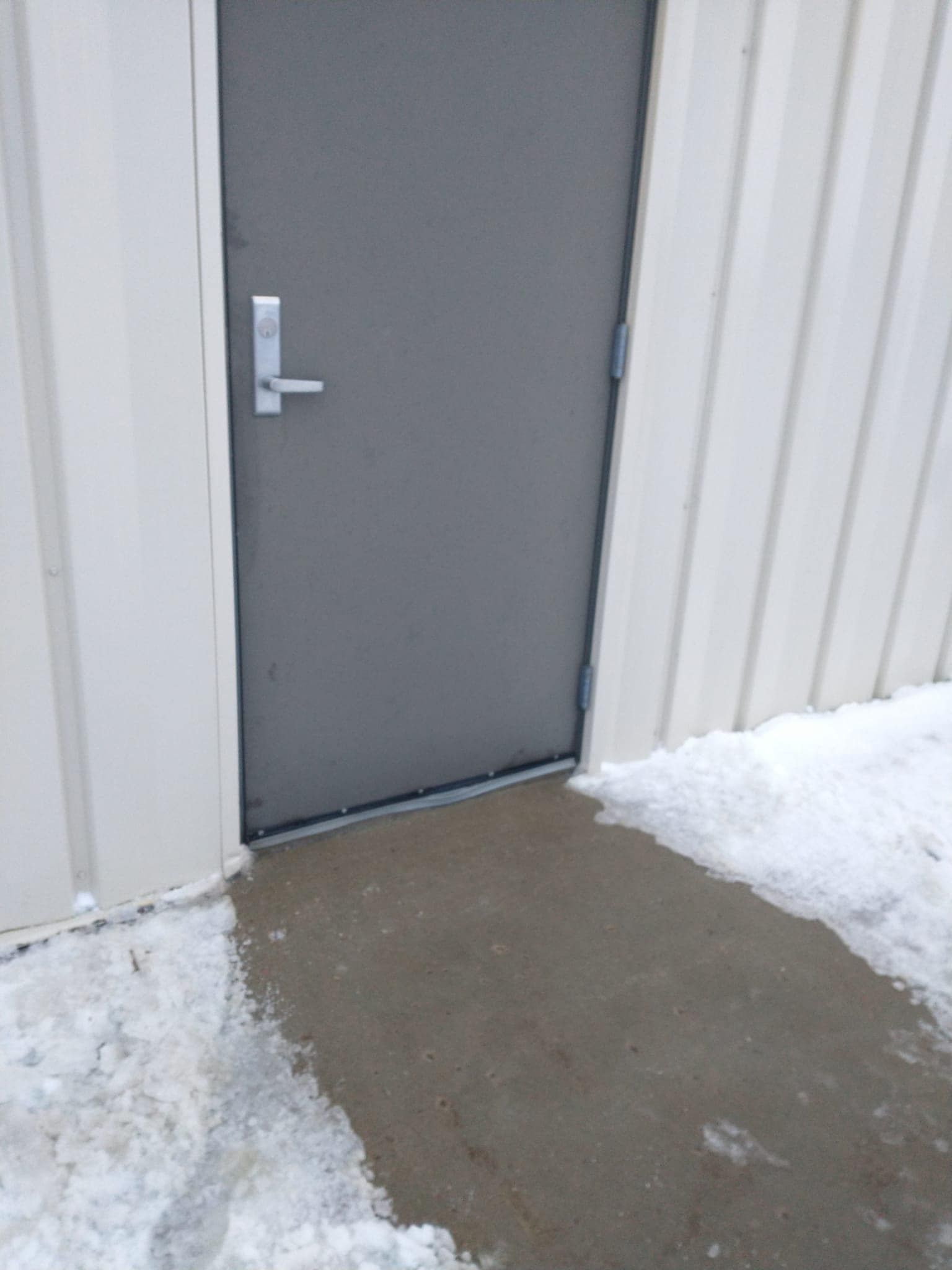 Gray door with silver handle in a white metal-sided building; concrete path with snow.