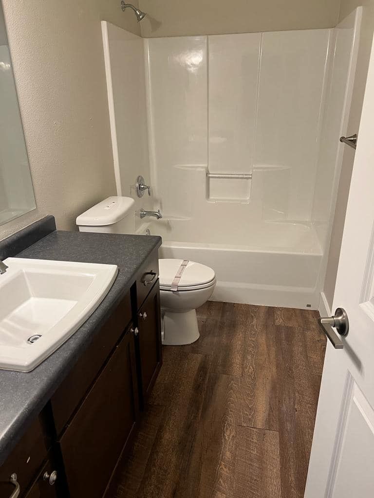 Bathroom with white tub/shower, toilet, dark wood vanity, and brown flooring.