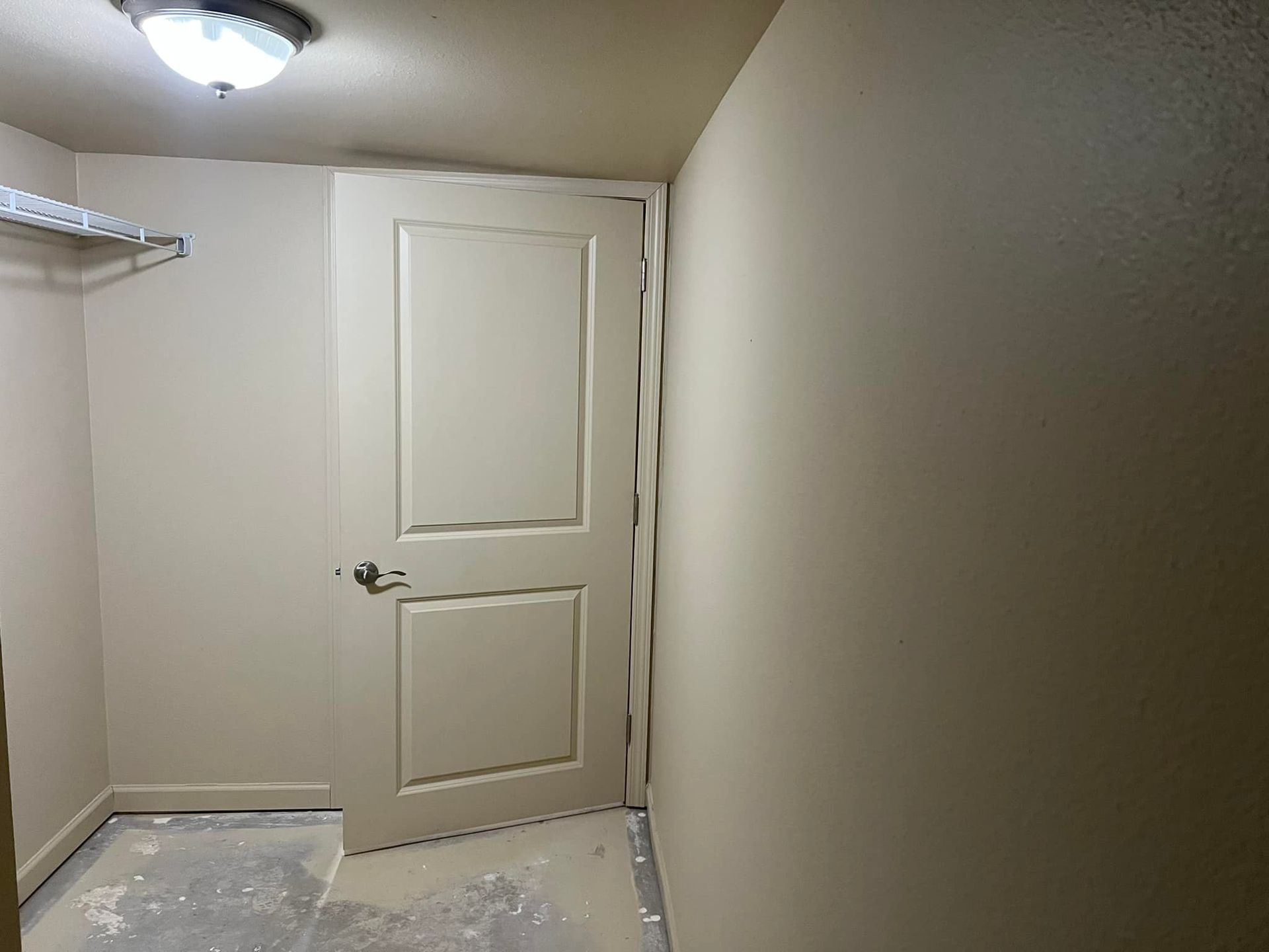 A small, empty room with a closed door, metal shelf, and a ceiling light. Walls are painted beige.