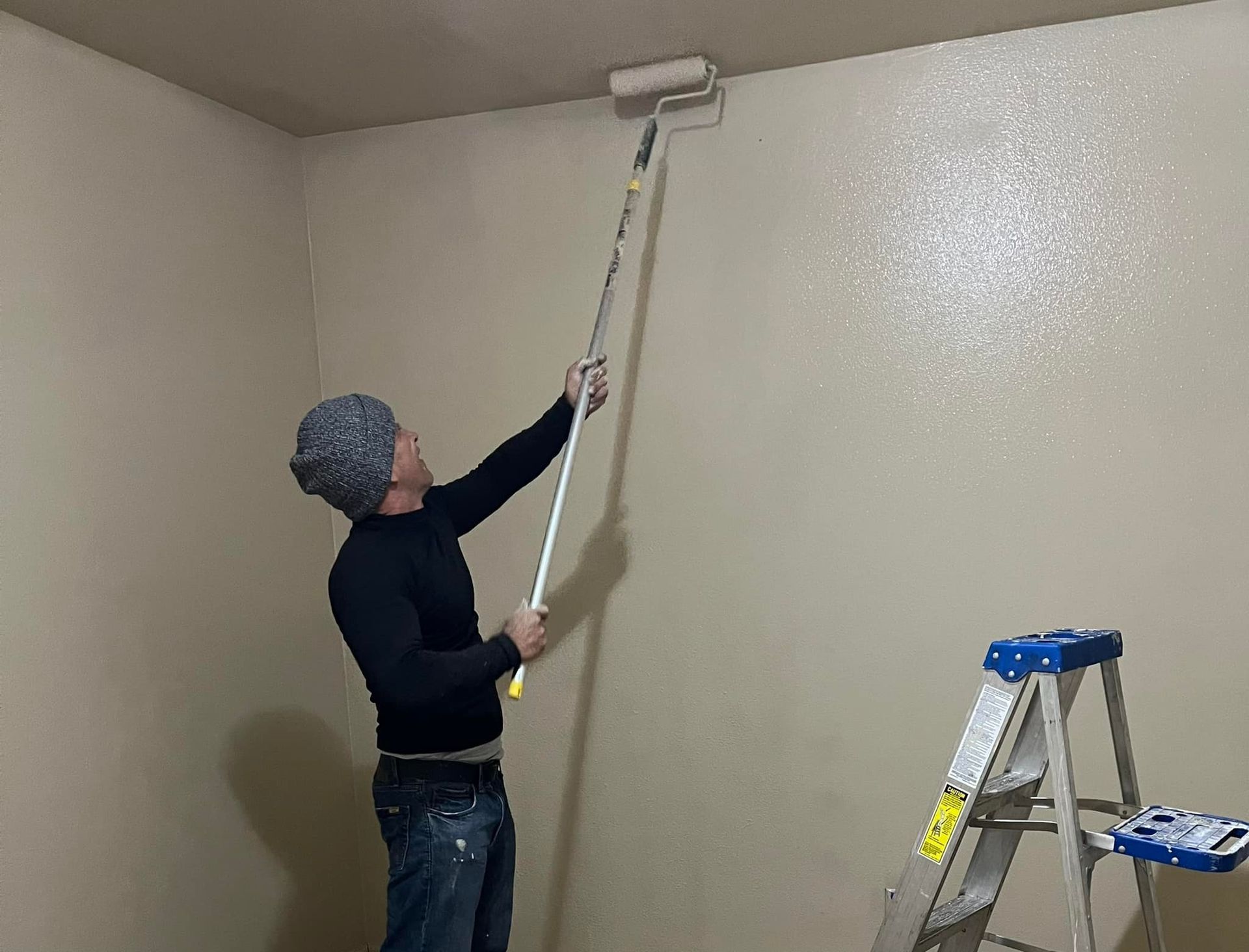 Person paints a wall with a roller. They are wearing a beanie, jeans, and a black shirt. A ladder is present.